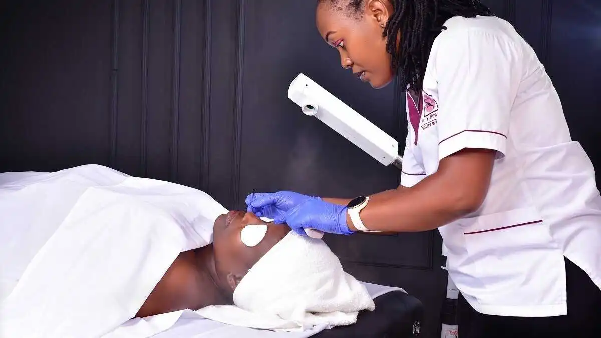 A beautician removes dead skin on the client's face. The client lays on the table covered in white sheet.