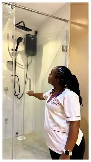A spa therapist pointing at a modern glass shower setup featuring dual rainfall and handheld shower heads, inside Viv’s In-Houz Spa treatment room at Kitisuru west mall