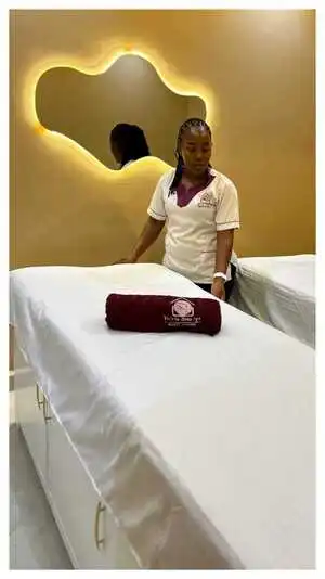 A spa therapist prepares a treatment bed in a warmly lit massage room at Viv’s In-Houz Spa studio at Kitisuru west mall, with a rolled towel branded with the spa logo and an artistic wall mirror in the background.