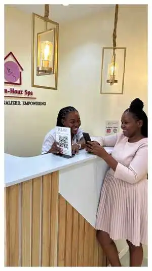 A smiling receptionist at Viv’s In-Houz Spa studio at Kitisuru west mall assists a client at the reception desk, showing a QR code for reviews, with elegant pendant lighting above.